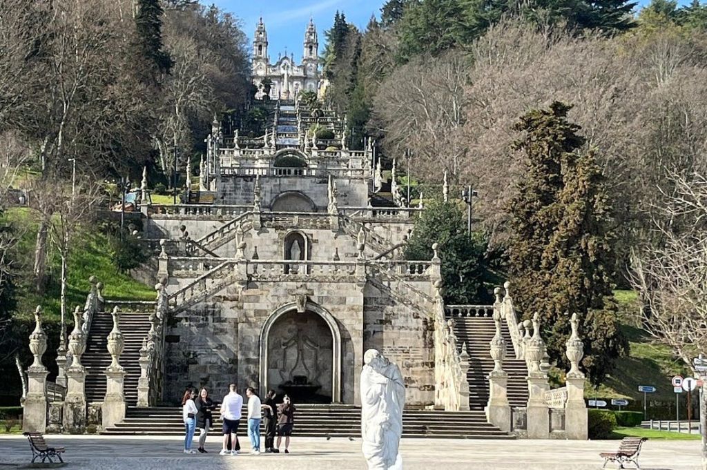 나무로 둘러싸인 화려한 건축물과 조각상이 있는 역사적인 건물로 이어지는 커다란 돌 계단 근처에 서 있는 사람들.