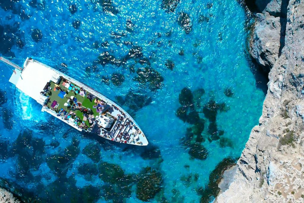 바위가 많은 해안선 근처의 맑고 푸른 바다에서 사람들이 갑판에 있는 흰색 보트의 공중 전망.