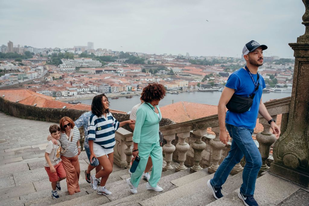 캐주얼한 옷을 입은 한 남성이 도시 풍경과 강을 배경으로 두 여성을 돌 계단으로 안내합니다.