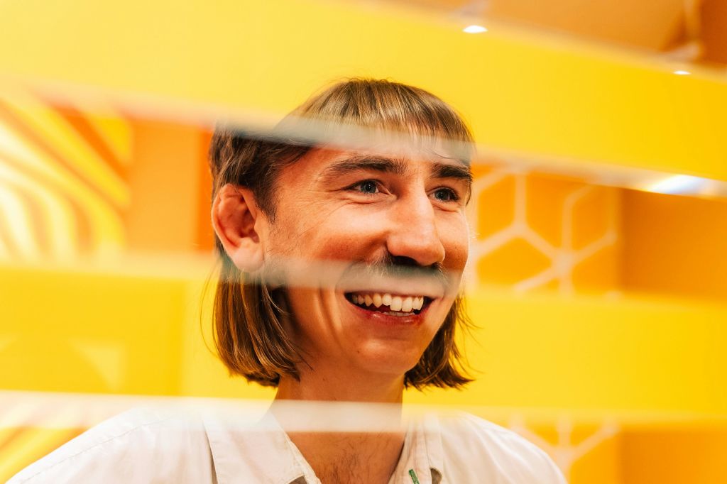 어깨 길이의 머리카락을 가진 웃는 사람이 육각형 패턴의 밝은 노란색 배경에 대해 수평 블라인드를 통해 보입니다.