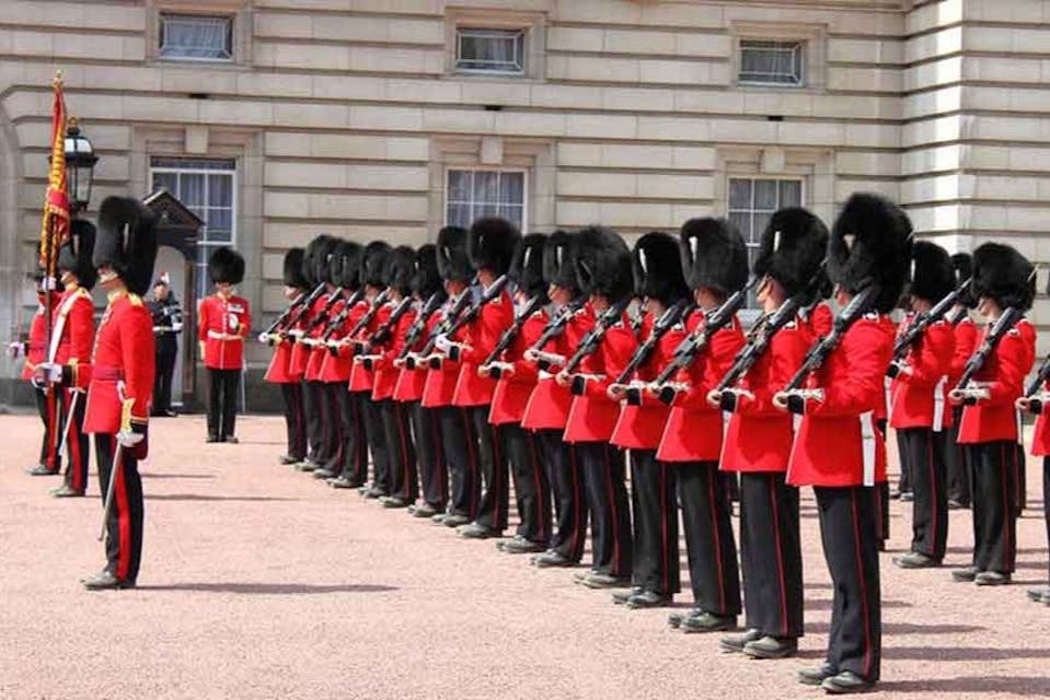 붉은 유니폼과 검은 곰 가죽 모자를 쓴 경비병들이 여러 개의 창문이 있는 석조 건물 바깥에 서 있습니다.
