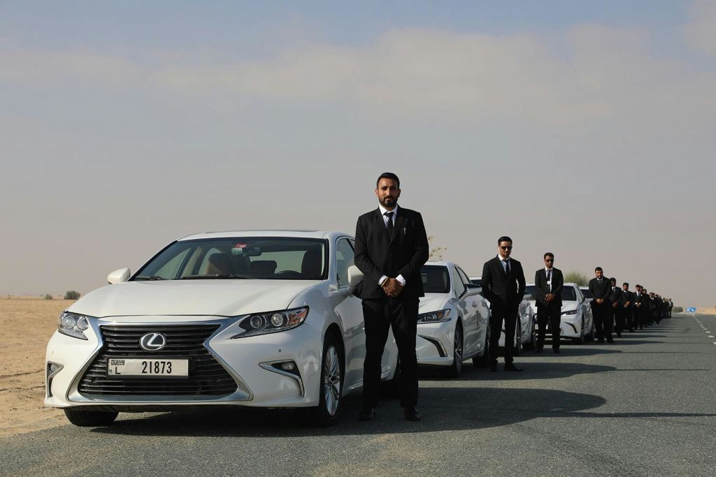 검은 양복을 입은 남성들이 부분적으로 흐린 하늘 아래 멀리까지 뻗어 있는 탁 트인 도로 위에 하얀 자동차 줄 옆에 서 있습니다.