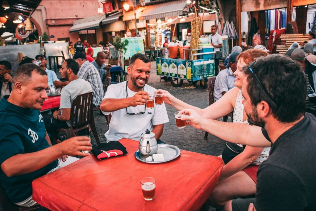 세 명의 남성이 야외 테이블에 앉아 상인과 가판대가 있는 분주한 시장에서 작은 차 한 잔과 함께 토스트를 하고 있습니다.