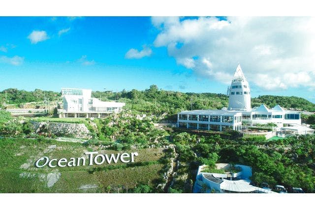 녹음으로 둘러싸인 현대적인 흰색 건물 단지로, 구름이 있는 푸른 하늘 아래 "오션 타워" 라는 이름의 높은 타워가 있습니다.