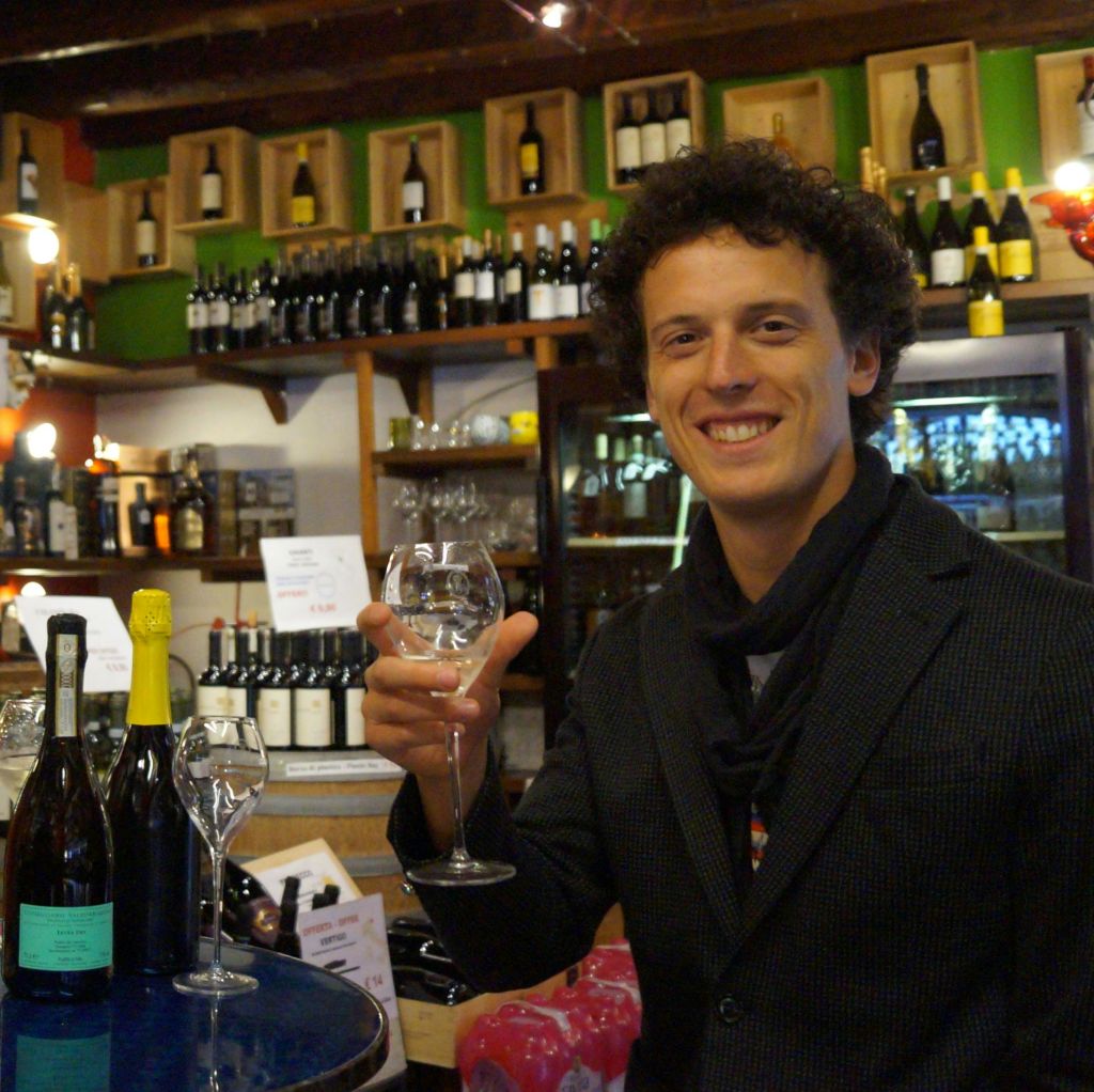 양복을 입은 남성이 와인잔을 들고 선반과 카운터에 놓인 병으로 가득 찬 와인숍에 서 있습니다.