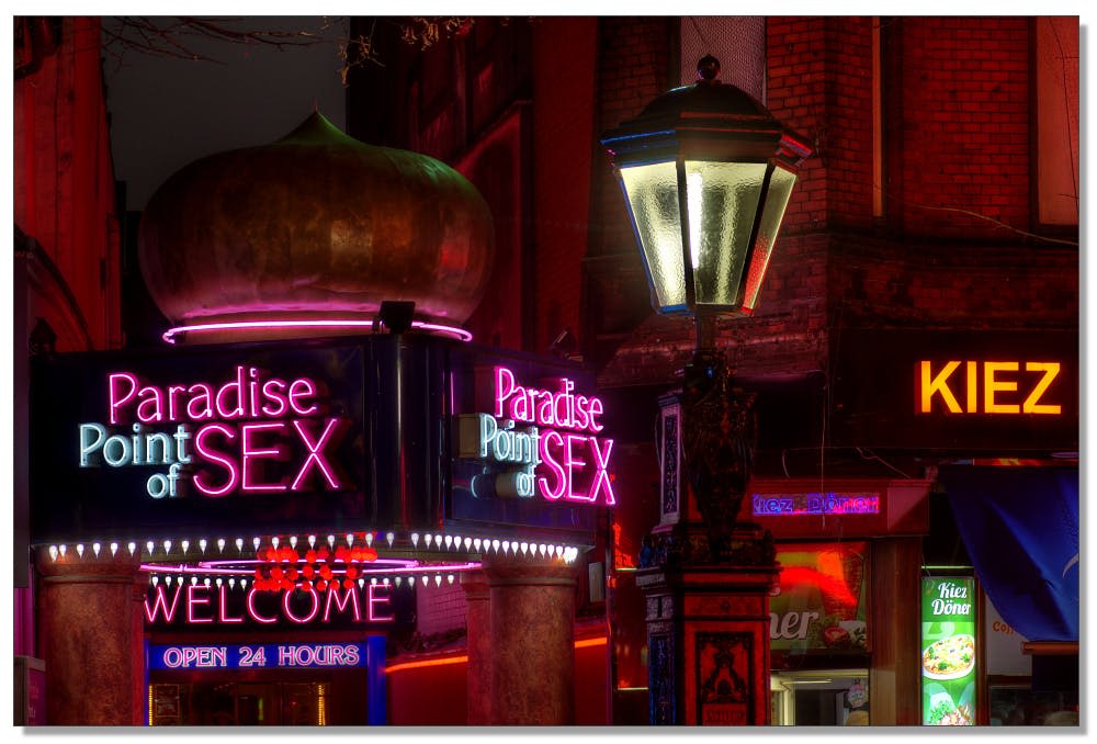 밤에 밝은 분홍색과 보라색 불빛이 비추는 "Paradise Point of SEX" 와 "WELCOME OPEN 24 HOURS" 의 네온 사인.
