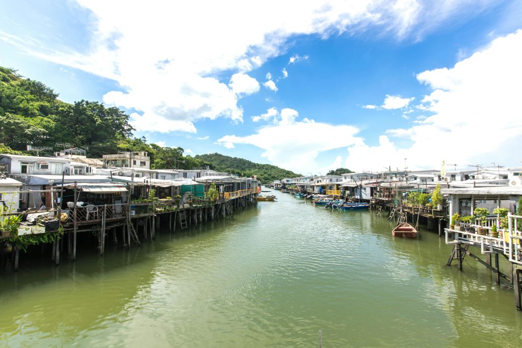 작은 보트가 정박되어 있고 언덕과 푸른 하늘을 배경으로 운하 양쪽에 줄지어 있는 철조 주택의 햇살이 내리쬐는 풍경입니다.