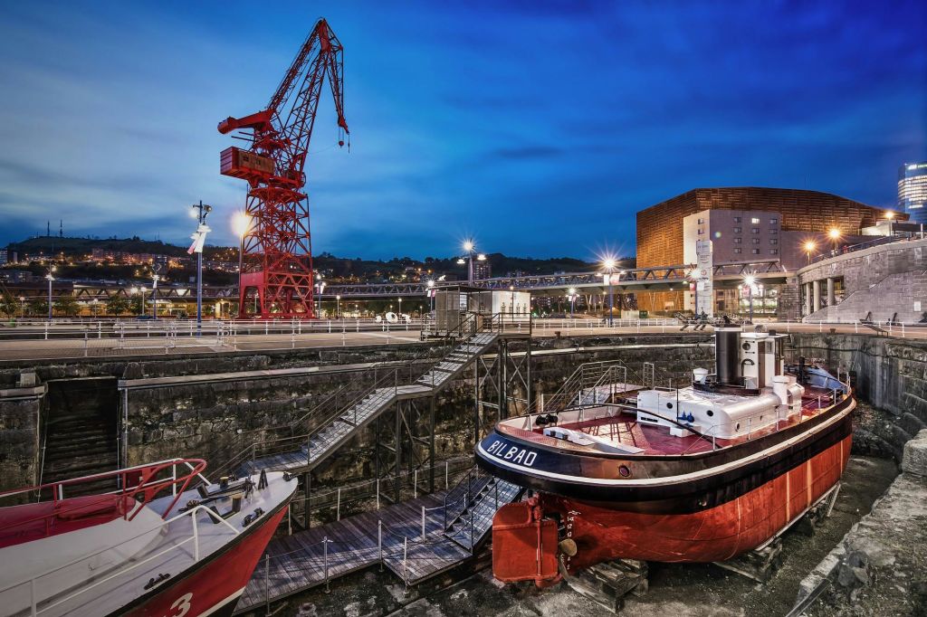 해질 무렵 보트, 빨간색 크레인, 다리, 가로등이 비추는 배경의 건물이 있는 선착장입니다.