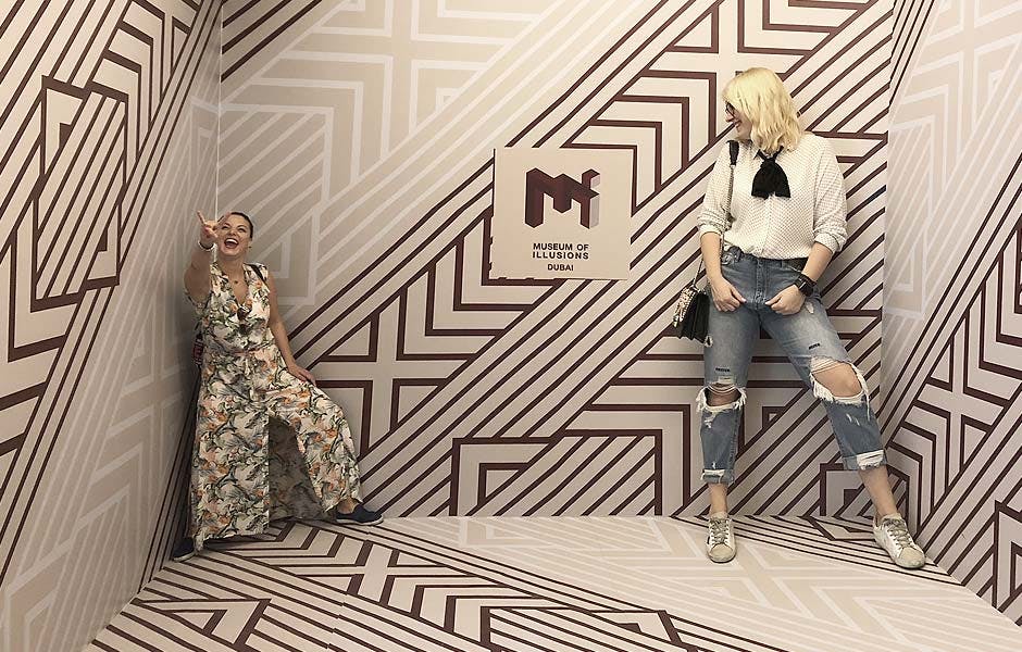 패턴이 있는 복장을 한 사람이 두바이 환상 박물관의 기하학적 벽 디자인이 있는 방에서 포즈를 취하며 미소를 짓고 포인트를 줍니다.