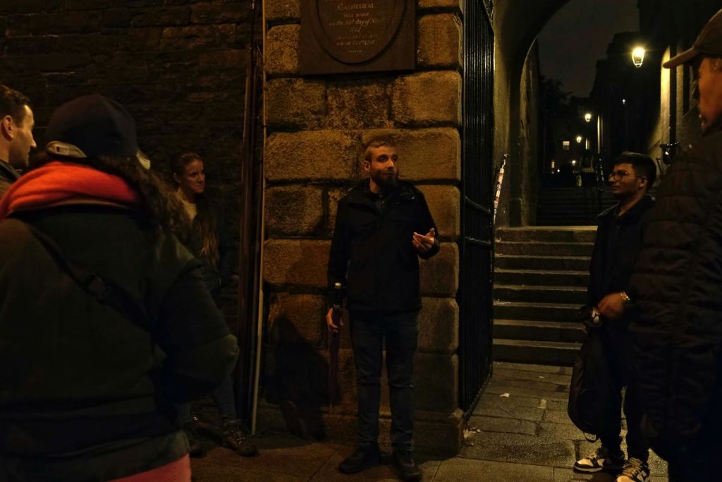 우산을 들고 말을 하는 수염을 기른 남자가 밤에 돌로 된 아치형 통로 옆에 서서 다른 두 남자가 듣고 있습니다.