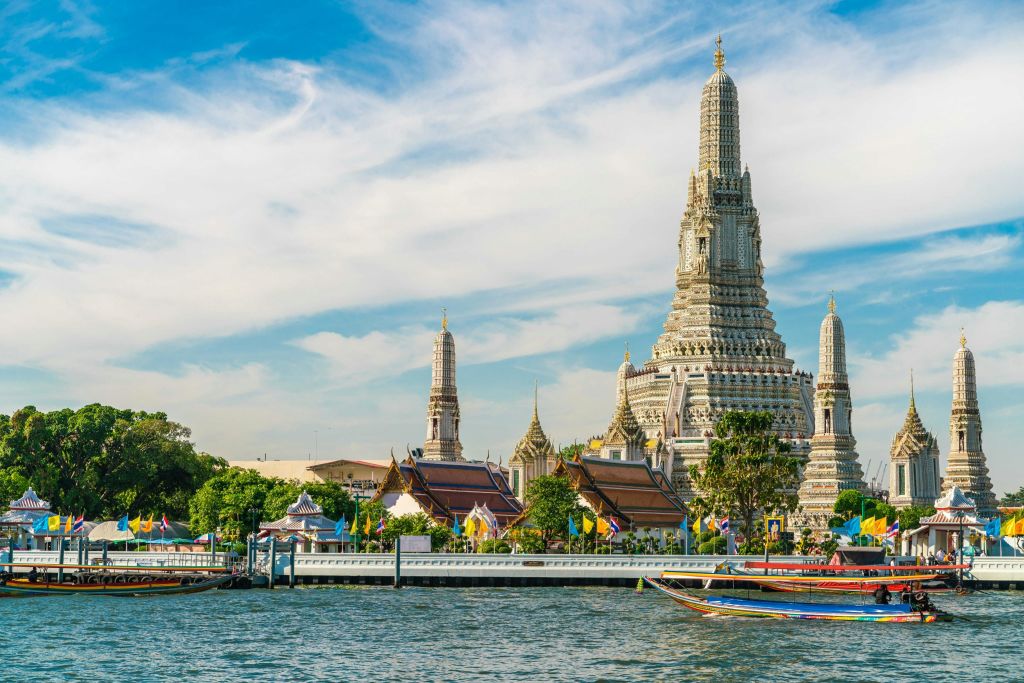 커다란 첨탑, 화려한 지붕이 있는 작은 건물, 화려한 롱테일 보트가 있는 방콕의 왓 아룬 사원의 강변 풍경.