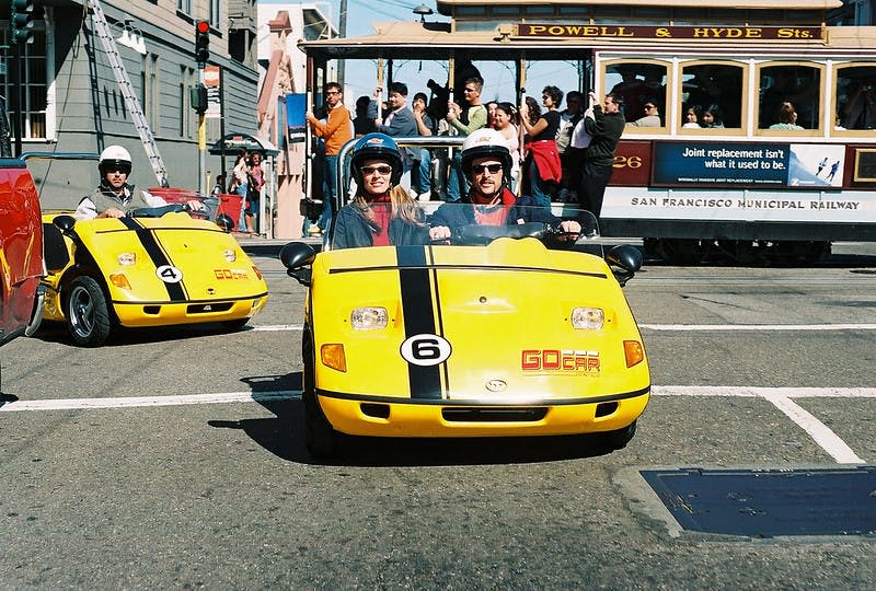 헬멧을 쓴 두 사람이 거리에서 노란색 고카를 타고 있습니다. 승객을 태운 파월 & 하이드 케이블카가 배경에 있습니다.
