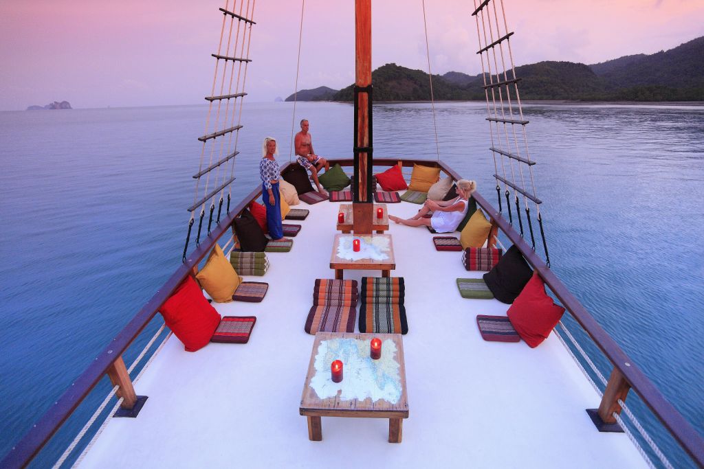 세 사람이 화려한 쿠션과 랜턴이 놓인 테이블이 있는 보트 데크에서 휴식을 취하고 있습니다. 잔잔한 물과 섬이 배경입니다.