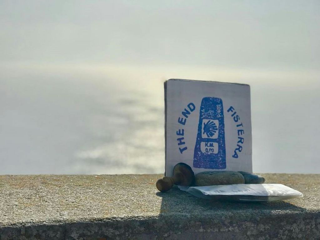 "The End Fisterra KM 0,0" 표시가 선반에 서 있으며, 바닷물과 햇빛이 배경으로 보입니다.