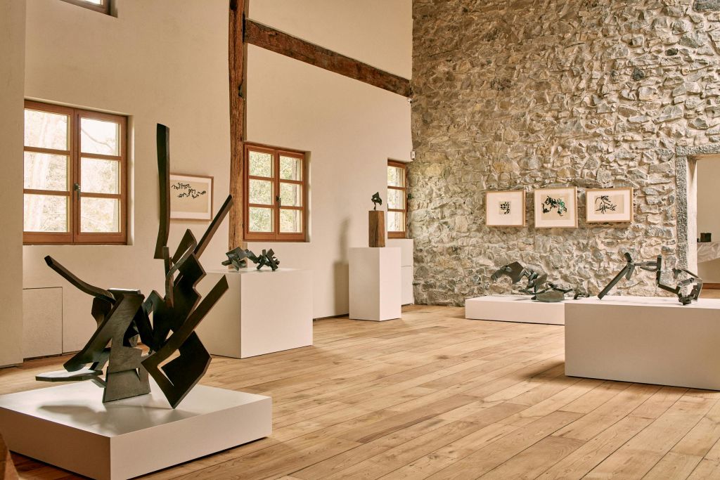 흰색 받침대에 현대적인 조각품, 돌 벽에 액자 예술품, 나무 바닥이 있는 갤러리 내부.