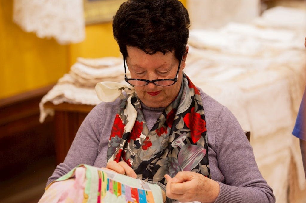 안경을 쓴 여성이 꽃무늬 스카프와 보라색 스웨터를 입고 화려한 패턴의 패브릭을 바느질합니다.