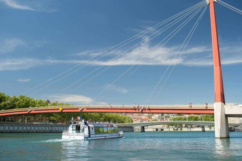 푸른 하늘 아래 건물과 나무를 배경으로 하얀 보트가 케이블이 달린 현수교 아래 강에 떠 있습니다.