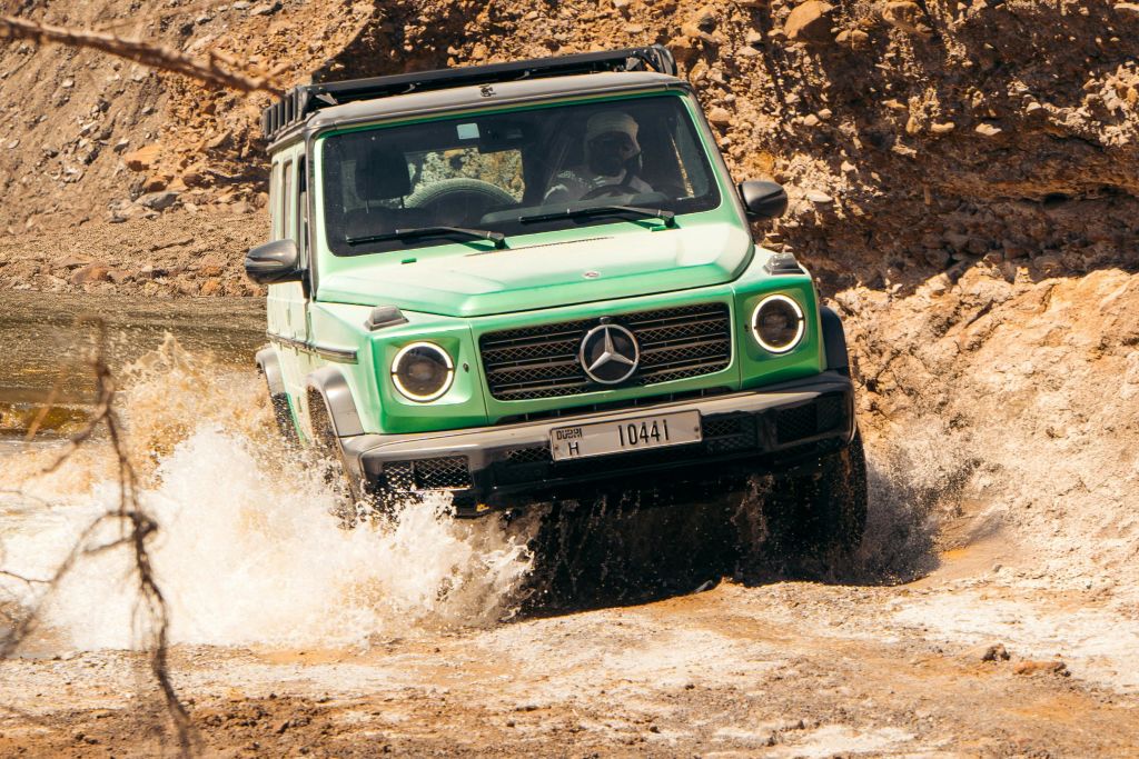 초록색 메르세데스-벤츠 SUV가 바위 지형의 물을 뚫고 운전하며 운전자가 내부를 볼 수 있도록 진흙탕을 튀깁니다.