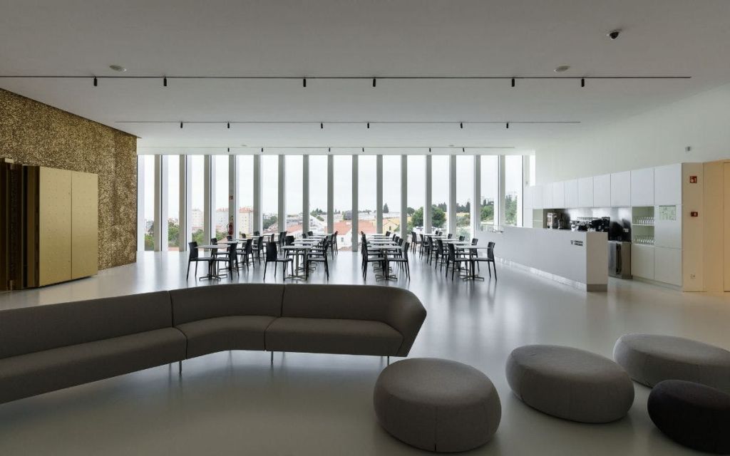 바닥에서 천장까지 이어지는 창문, 빈 테이블과 의자, 둥근 오토만이 있는 곡선형 소파가 있는 넓고 현대적인 카페테리아입니다.