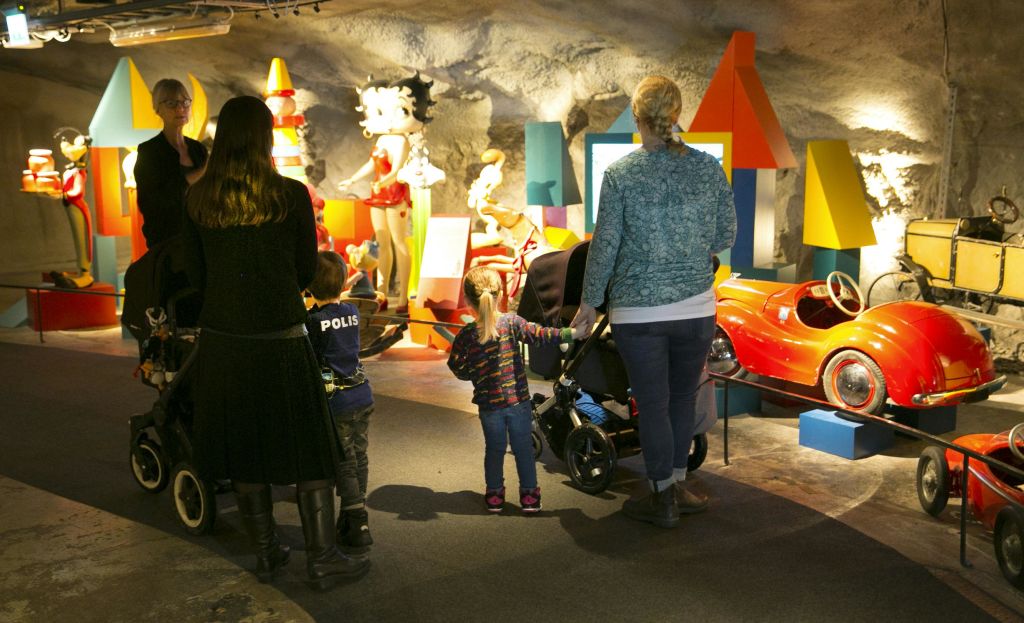 다채로운 조각상과 전광판이 있는 방에 어른과 아이들이 있고, 일부 유모차가 있습니다. 씬에 조명이 잘 들어옵니다.