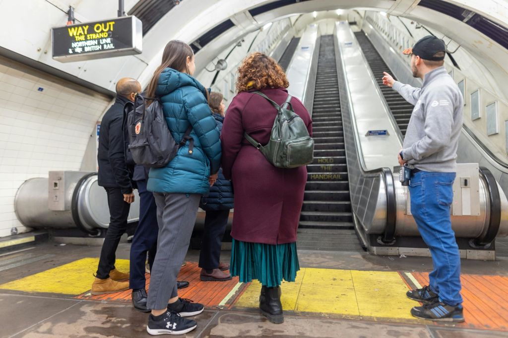 위에 표지판이 보이는 지하철이나 기차역의 에스컬레이터 밑에 서 있는 사람들.