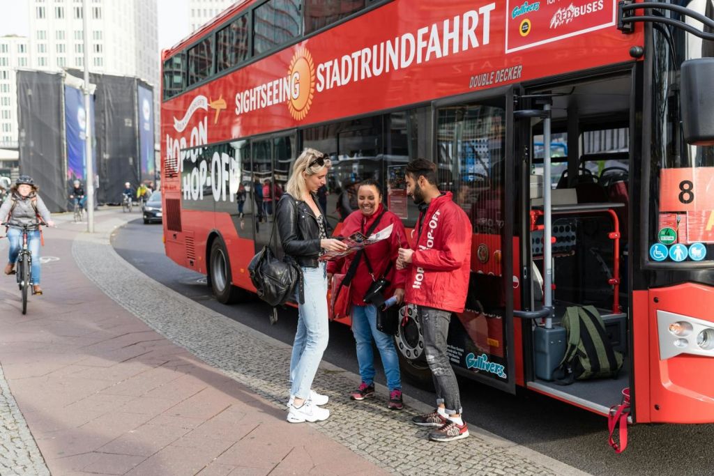 빨간색 2층 관광 버스 옆에 세 사람이 서 있습니다. 한 사람이 지도를 들고 직원 2명과 이야기를 나누고 있습니다.