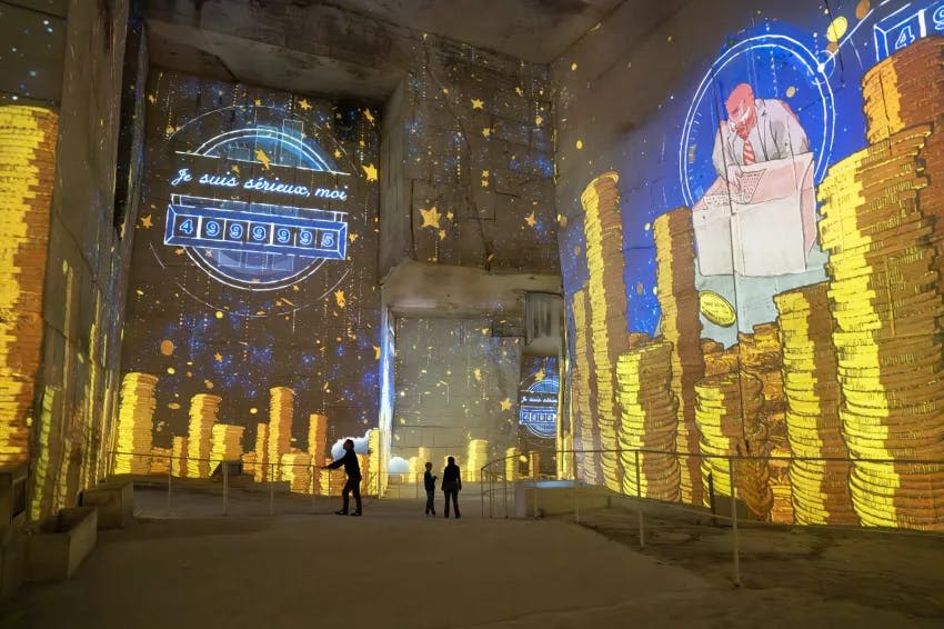 동굴 같은 공간에 금화, 별, 한 남자가 책상에 투영된 몰입형 예술 작품이 설치되어 있습니다.