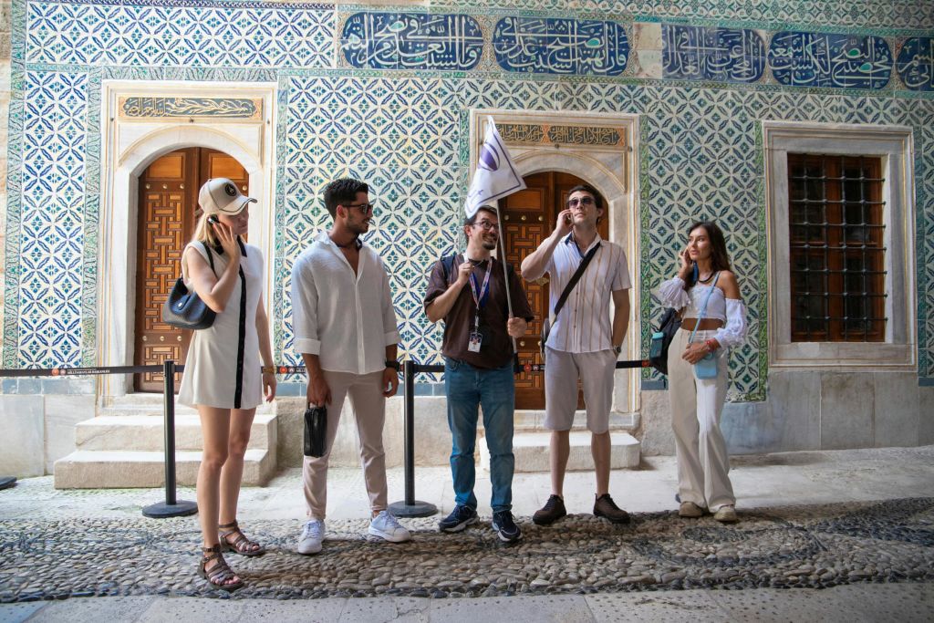 아랍 문자가 쓰여진 무늬가 있는 벽 앞에 네 사람이 서 있습니다. 한 사람이 깃발을 들고 있어 투어를 진행할 수 있습니다.