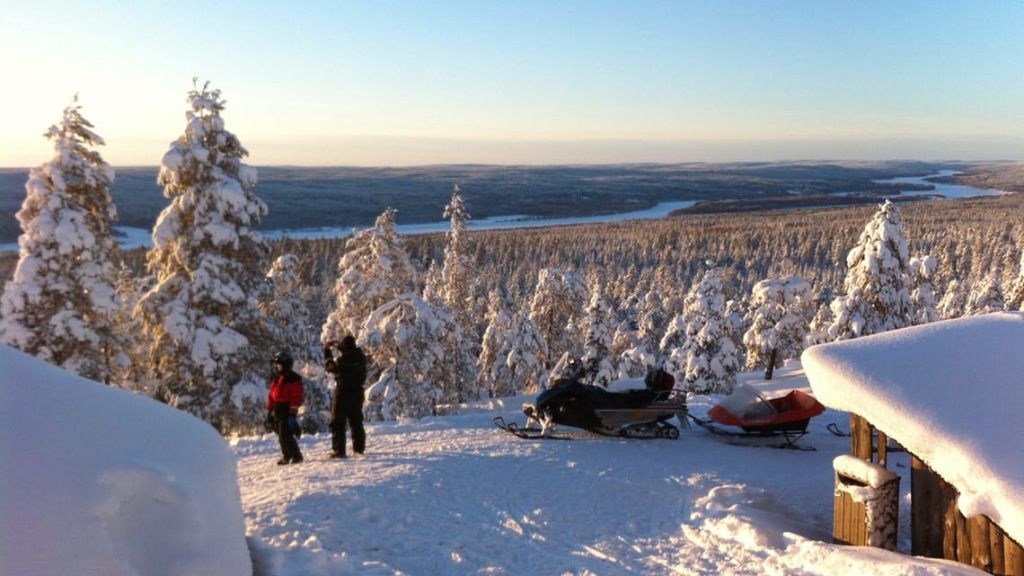 맑은 하늘 아래 눈 덮인 광활한 숲이 내려다보이는 눈 덮인 언덕에 겨울 옷을 입은 두 사람이 스노모빌 근처에 서 있습니다.