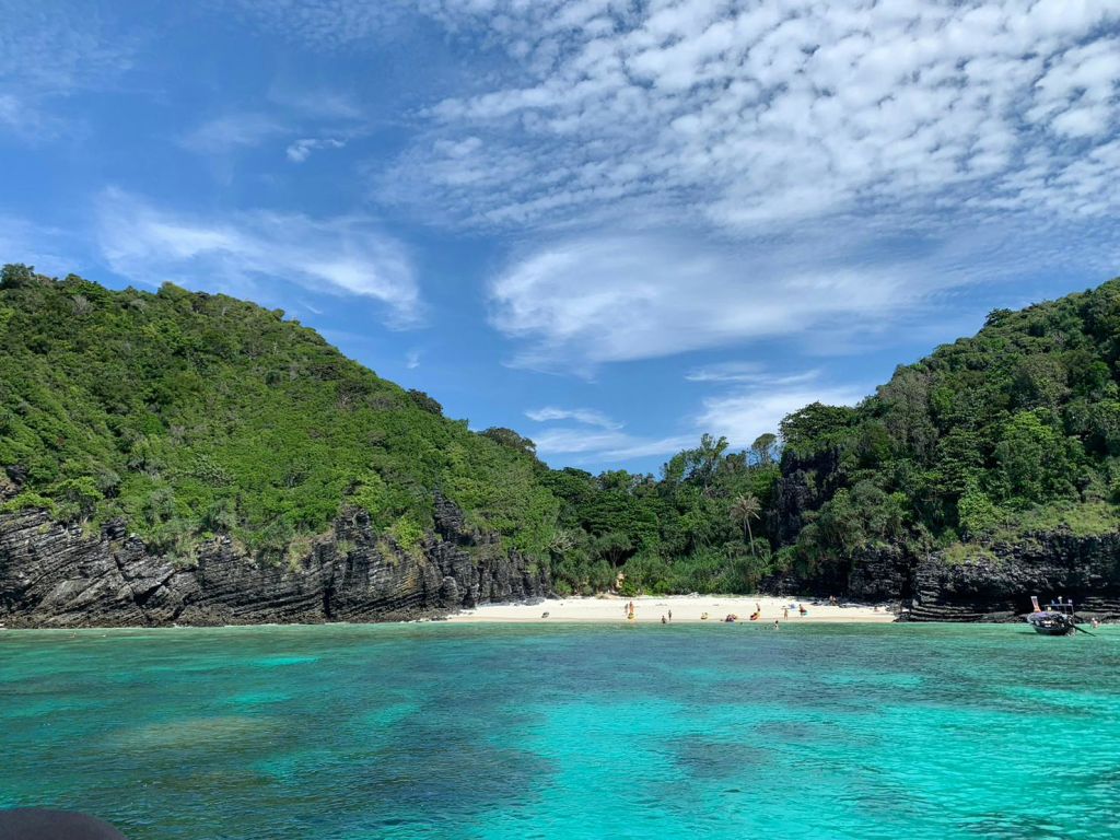 푸른 언덕이 우거진 깨끗한 해변, 청록색의 맑은 물, 부분적으로 흐린 하늘 아래에서 휴식을 취하는 소수의 사람들.