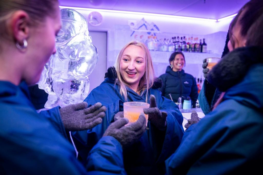 겨울 재킷을 입은 사람들은 얼음 조각과 웃는 바텐더를 배경으로 얼음으로 만든 바에서 음료를 즐깁니다.