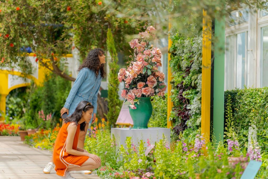 다양한 식물과 다채로운 꽃이 만발한 활기차고 울창한 정원에서 두 여성이 커다란 분홍색 꽃병을 감상하고 있습니다.