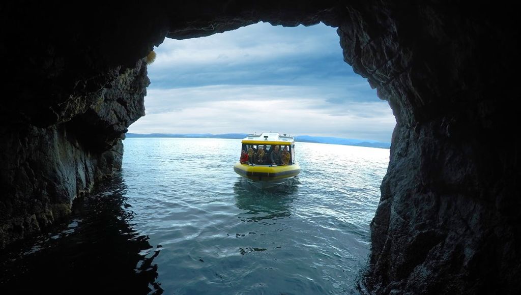 구름이 낀 하늘을 배경으로 잔잔한 푸른 물 위에 떠 있는 동굴에서 사람들이 타고 있는 노란 보트가 보입니다.