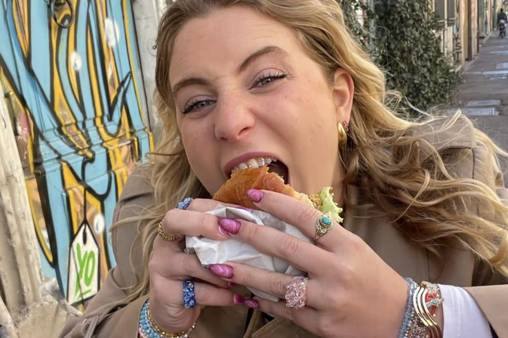 그래피티 태그가 달린 벽 앞에서 카메라 렌즈를 바라보며 샌드위치를 먹는 여성