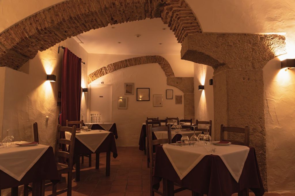 나무 테이블과 의자, 흰색 식탁보, 아치, 액자 사진, 테라코타 타일 바닥이 있는 조명이 희미한 레스토랑 내부.