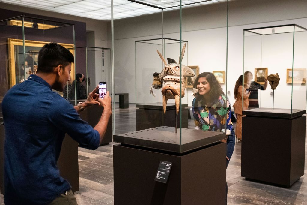 박물관의 유리 케이스에 전시된 부족 마스크를 보며 미소를 짓고 감탄하는 여성과 다른 사람이 사진을 찍고 있습니다.