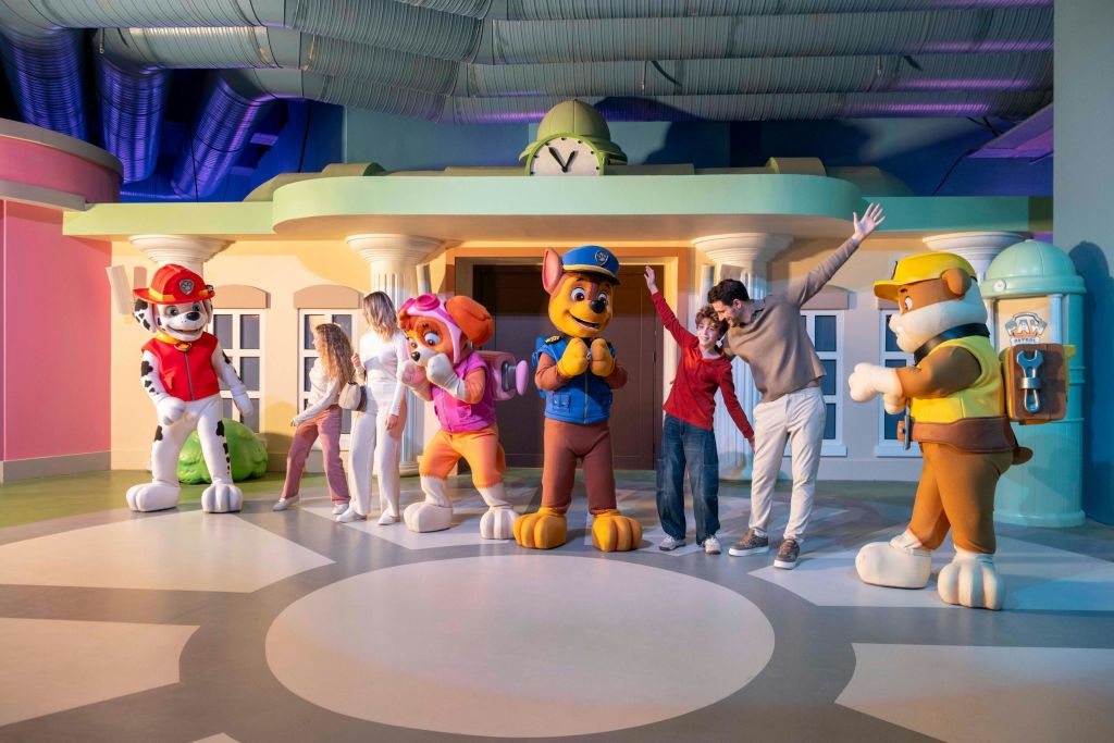 만화 마을을 닮은 다채로운 실내 환경에서 어린이 공연의 캐릭터와 함께 포즈를 취하는 사람들.