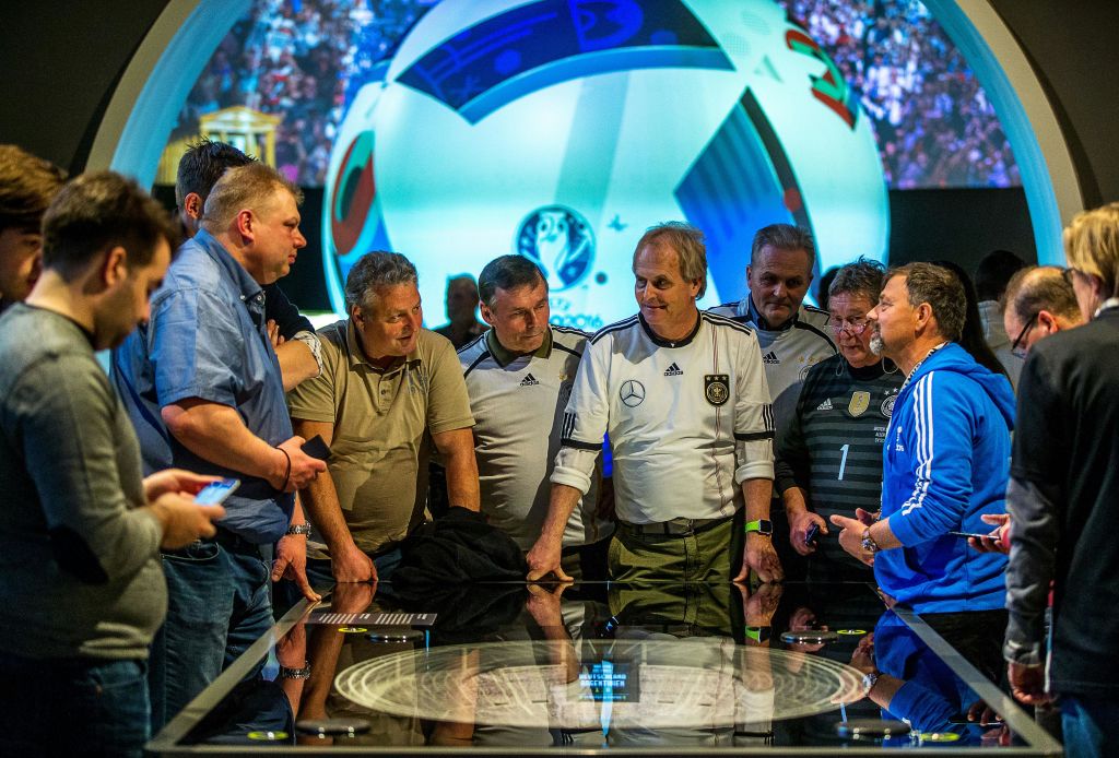 스포츠 유니폼을 입은 남성들이 커다란 축구공과 경기장 장면을 배경으로 인터랙티브 디스플레이에 모입니다.
