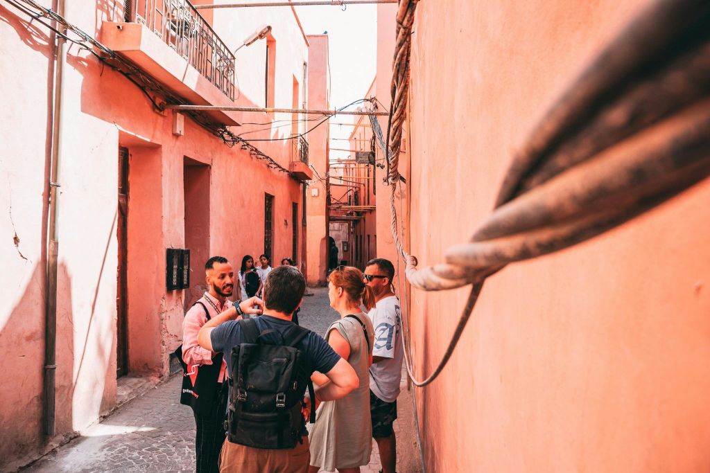 오후 햇살 아래 분홍색 벽과 노출된 전선, 작은 창문이 있는 좁은 골목길에서 한 무리의 관광객이 대화를 나누고 있습니다.