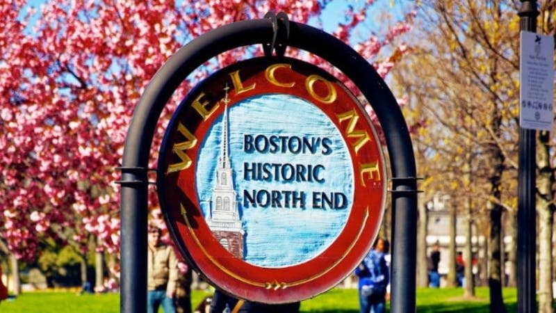 "Welcome Boston's Historic North End" 라는 글자와 봄 단풍을 배경으로 한 교회 첨탑 이미지가 있는 둥근 표지판.