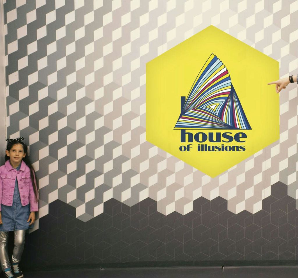 분홍색 재킷을 입은 어린이가 6각형 "환상의 집" 간판이 있는 무늬가 있는 벽에 서 있습니다.