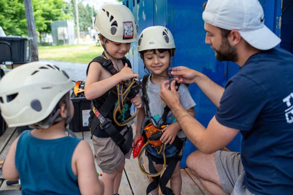 헬멧과 하네스를 착용한 두 어린이가 파란색 수납 공간 근처에서 지퍼 라이닝 활동을 준비할 수 있도록 성인이 도와줍니다.