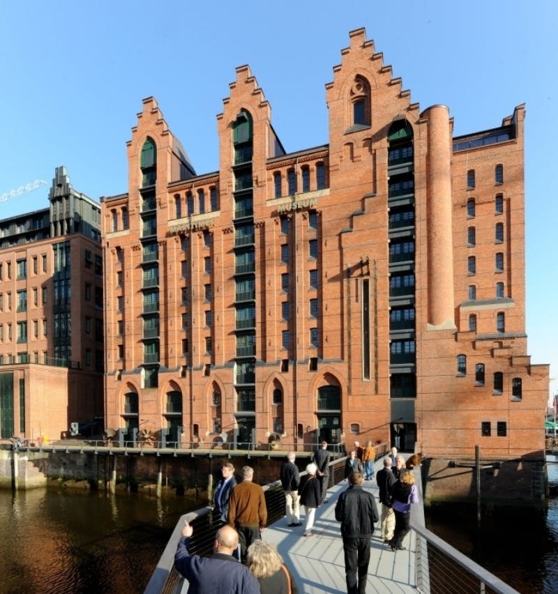 화창한 날에 고딕 양식의 건축물이 있는 커다란 붉은 벽돌 건물을 향해 다리를 걷는 사람들.