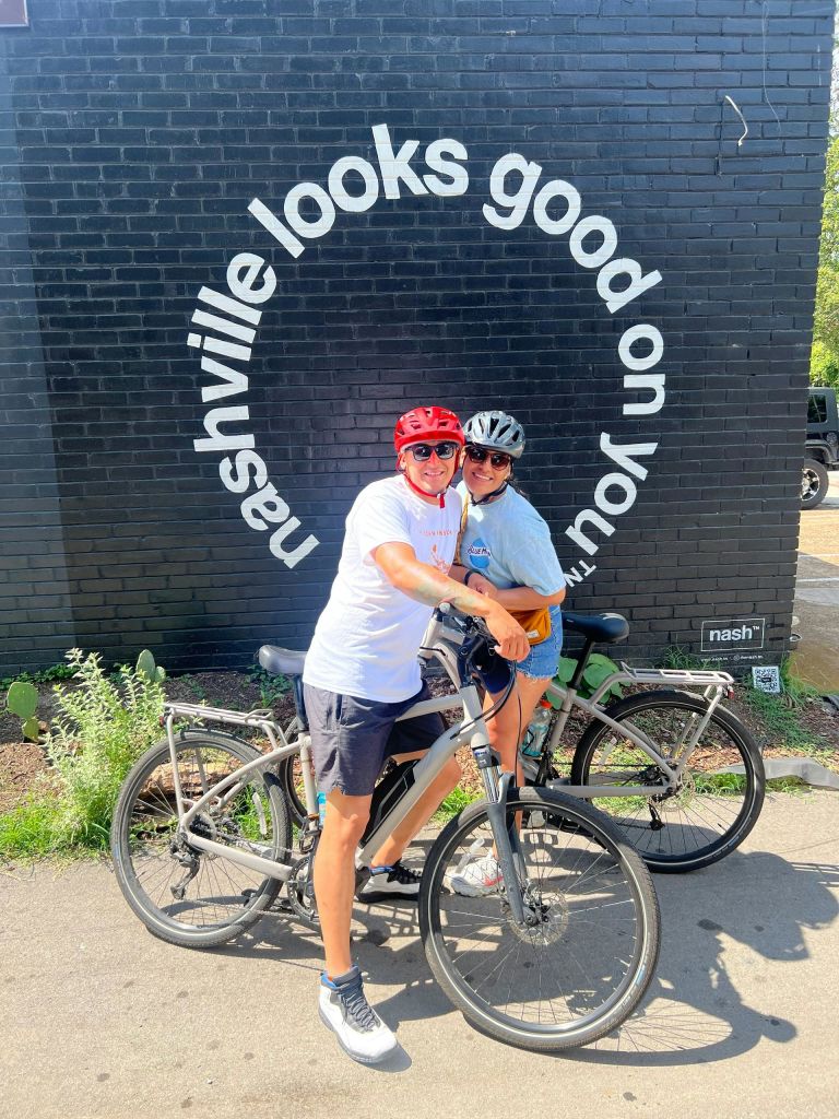 자전거 장비를 착용한 두 사람이 자전거를 타고 벽화 앞에 서서 '내슈빌이 당신에게 잘 어울립니다.' 라고 적혀 있습니다.