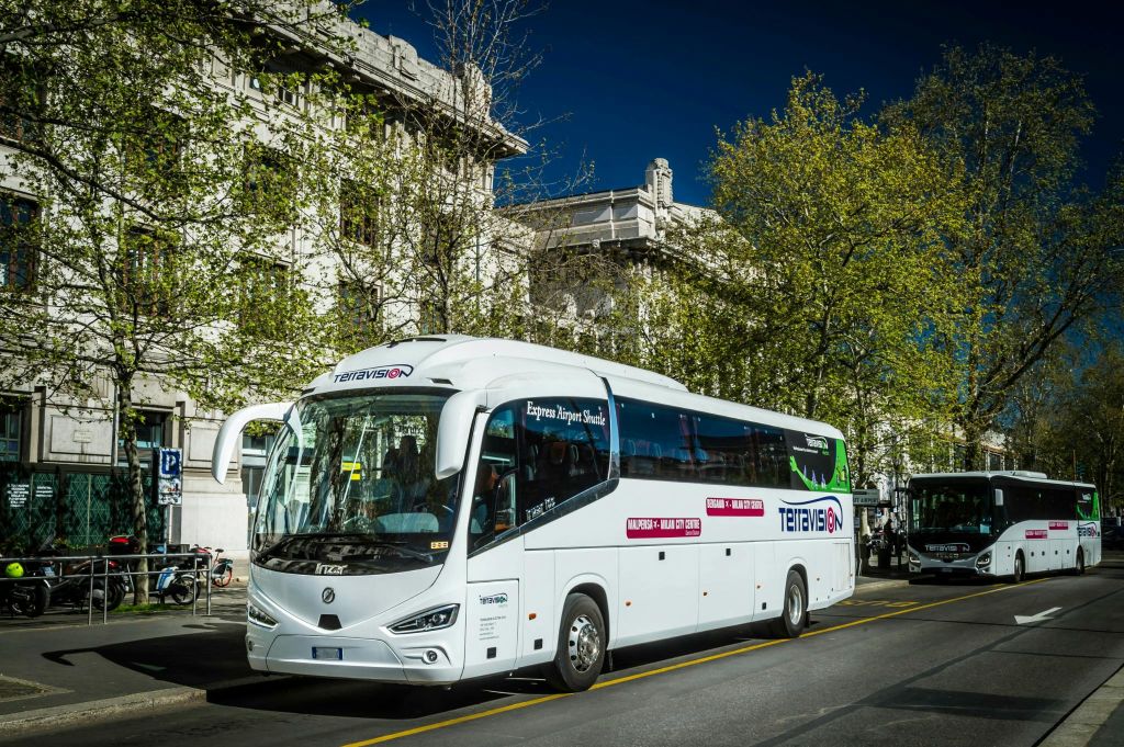 나무가 우거진 거리에 흰색 텔레비전 브랜드 버스가 주차되어 있으며, 역사적인 건물이 배경으로 있습니다.