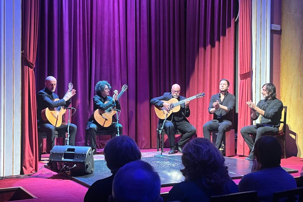 4명의 뮤지션이 무대에서 기타를 치고 박수를 치며 붉은 커튼 앞에 앉아 관객을 지켜보고 있습니다.