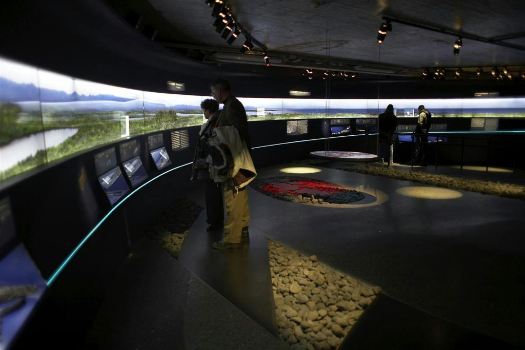 사람들은 원형 디스플레이와 오버헤드 조명이 있는 희미한 조명의 박물관에서 스크린과 안내 패널이 있는 전시물을 봅니다.