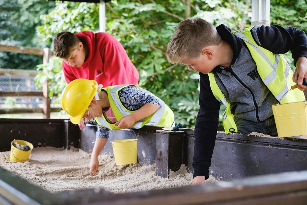 녹음으로 둘러싸인 캐노피 아래에서 노란 양동이가 있는 모래를 파고 있는 노란 조끼를 입은 어린이.