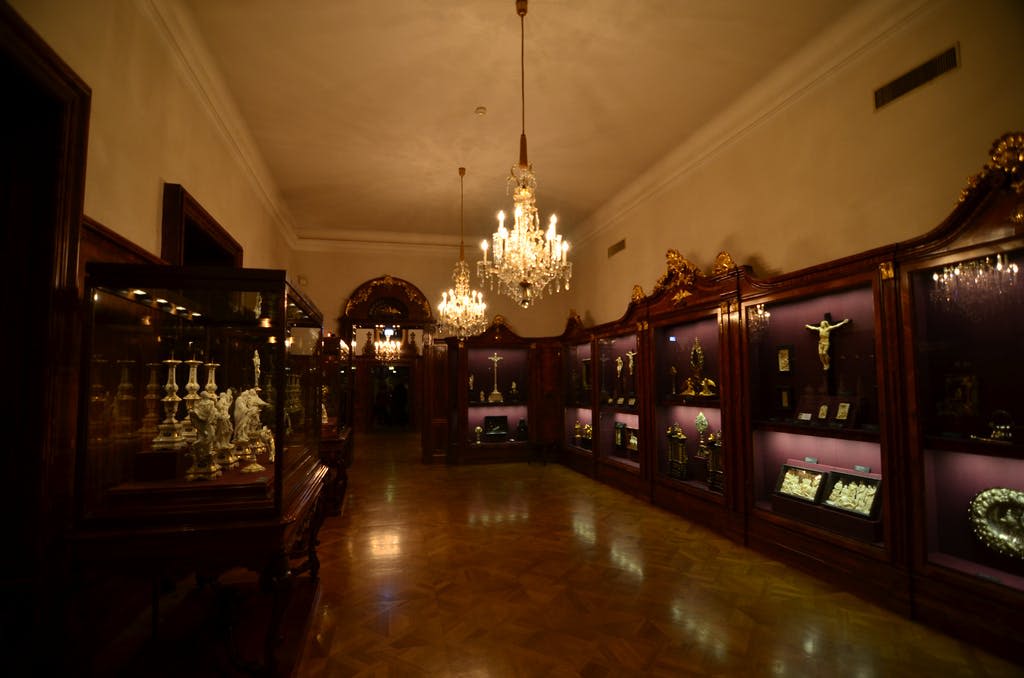 샹들리에가 있는 화려한 객실로, 보라색 조명이 있는 나무 진열 케이스에 종교 유물을 전시합니다. 광택이 나는 나무 바닥.