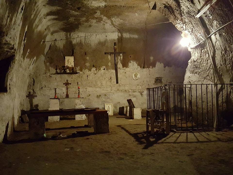 석조 벤치, 여러 개의 십자가, 촛불, 종교 아이콘, 오른쪽에 울타리가 쳐진 공간이 있는 조명이 희미한 지하 예배당입니다.
